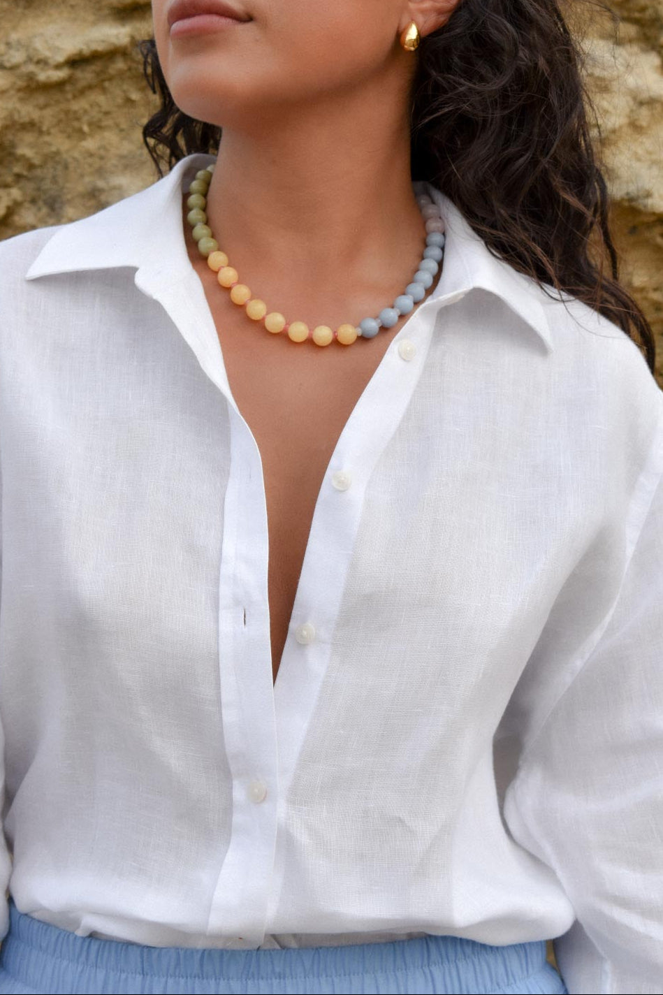 Woman wearing a white shirt with a necklace and earrings against a stone wall.